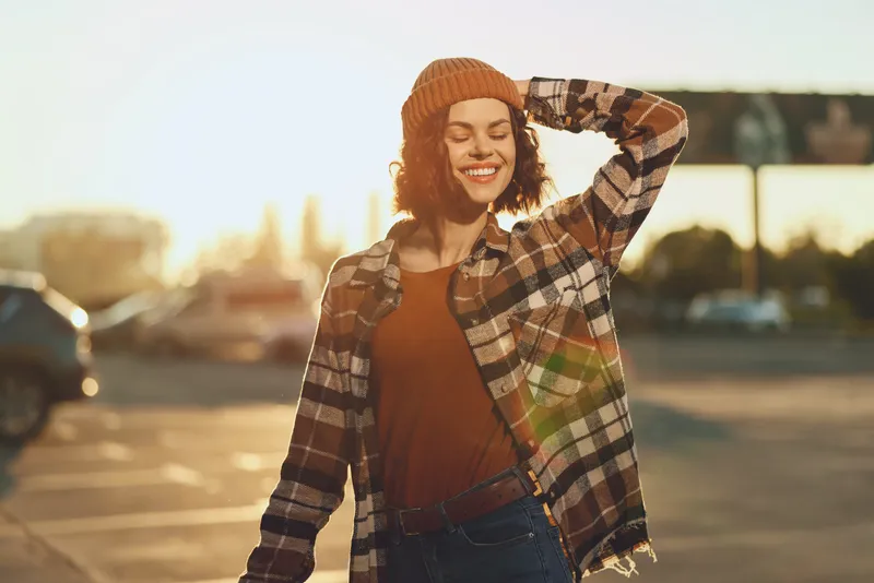 Woman with smile and eyes closed in golden hour glow, candid beanie and plaid outfit in urban parking lot under warm sunlight, authenticity, candid lifestyle, mindful living and emotional
