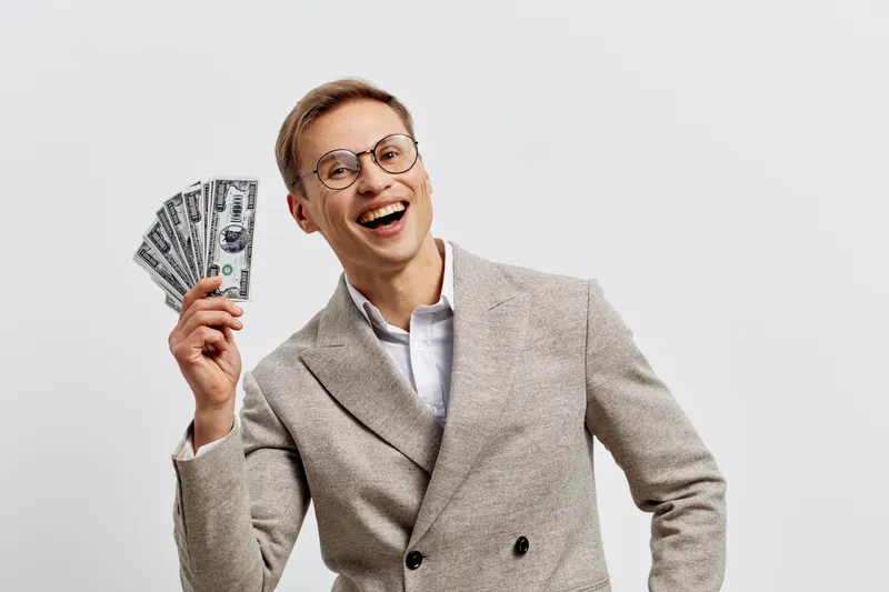 Happy young man hold fan of dollar bills wear glasses beige blazer smile isolated on white background digital nomad freelance success concept