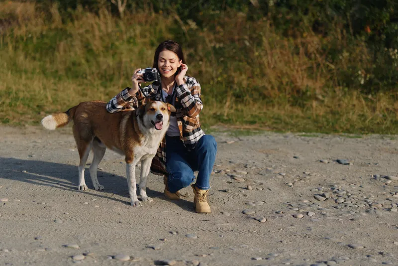 Woman kneels on a gravel beach, taking photos of a friendly dog with a camera, outdoor scene, casual outfit, bright daytime light, a playful moment captured on the shore.