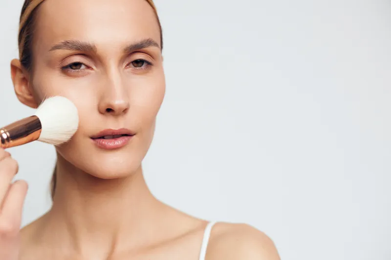 Closeup portrait of young woman applying makeup with brush on clean face, soft natural light, neutral background, minimalistic beauty routine, calm expression, skincare focus