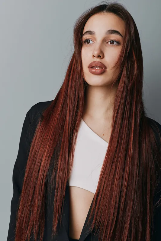 Young woman with long straight red hair wearing black jacket and white top standing against gray background looking away with serious expression and natural makeup.