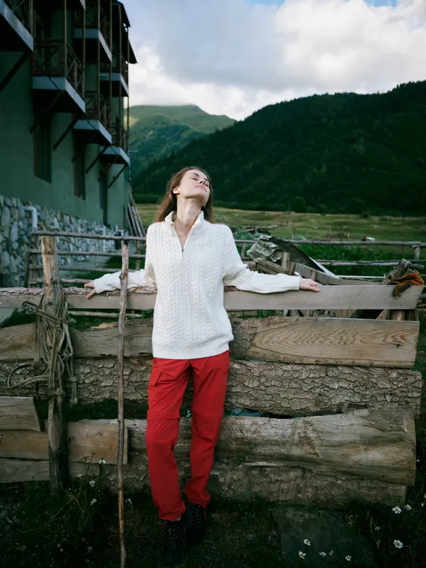 Woman stands outdoors by a rustic fence, looking up toward the mountains, wearing white blouse and red pants, capturing a moment of freedom in nature.