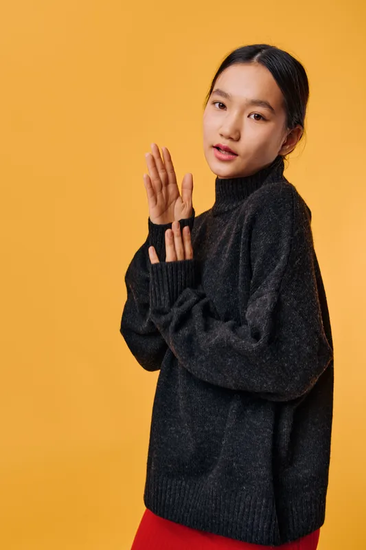 Young woman with long black hair wearing black sweater standing on orange background posing with hands together looking at camera with natural makeup and casual style.