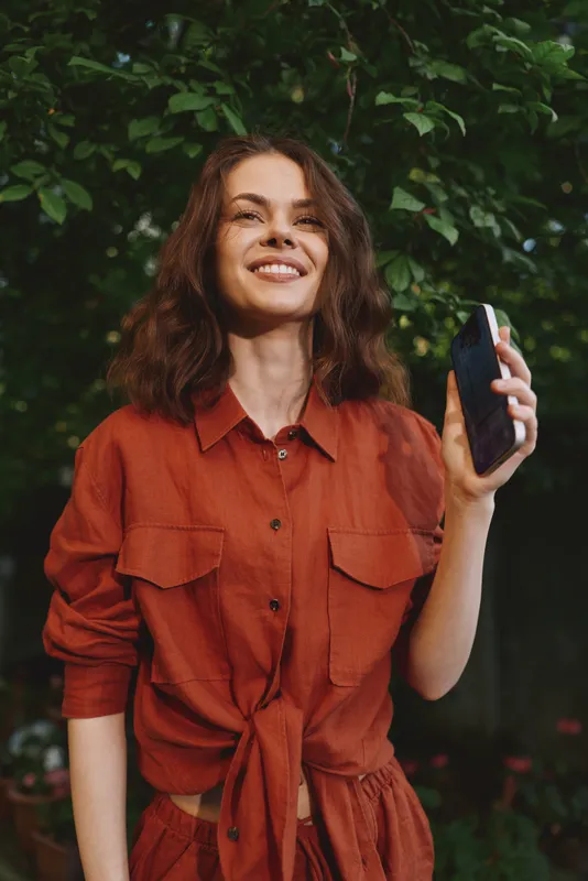 Young woman with wavy brown hair wears an orange outfit and smiles outdoors while holding a smartphone, capturing a candid moment in a sunlit garden
