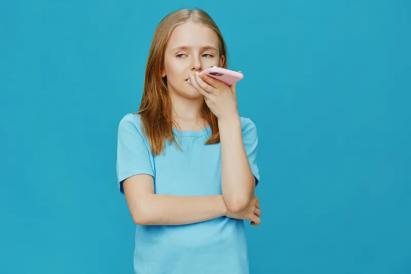 Young girl wearing a blue t shirt, looking pensive while holding a phone against a bright turquoise background Emphasizing emotions of contemplation and connection