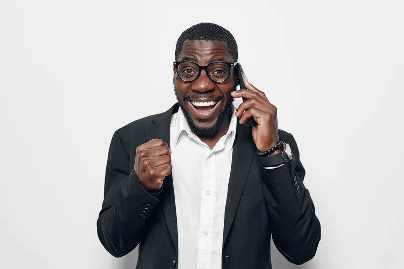 African American man with dark skin wearing glasses and suit talking on phone with excited expression and clenched fist for success and positive news on white background.