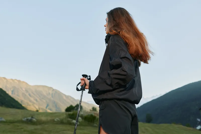 A lone traveler stands outdoors in a mountainous valley, gazing toward distant peaks. She holds a trekking pole, wearing a dark jacket, emanating calm focus and adventurous energy.