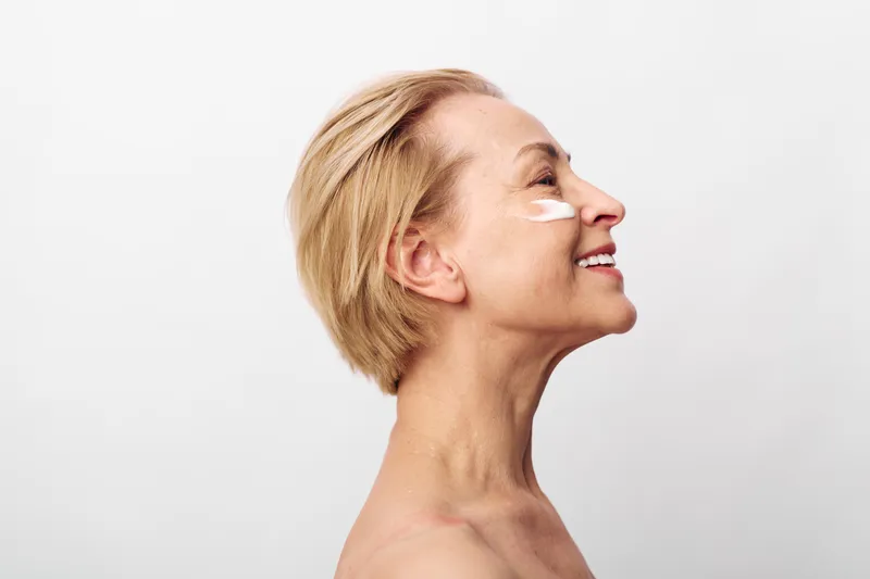 Smiling mature woman with short blonde hair applies white facial cream on cheek. Side profile on white background. Concept for skincare, antiaging, beauty routine, and self care.