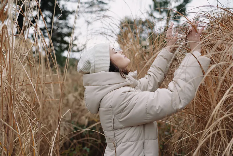 Outdoor winter scene with a woman in a light coat reaching through tall grass in an open field, conveying calm, nature, and a contemplative moment
