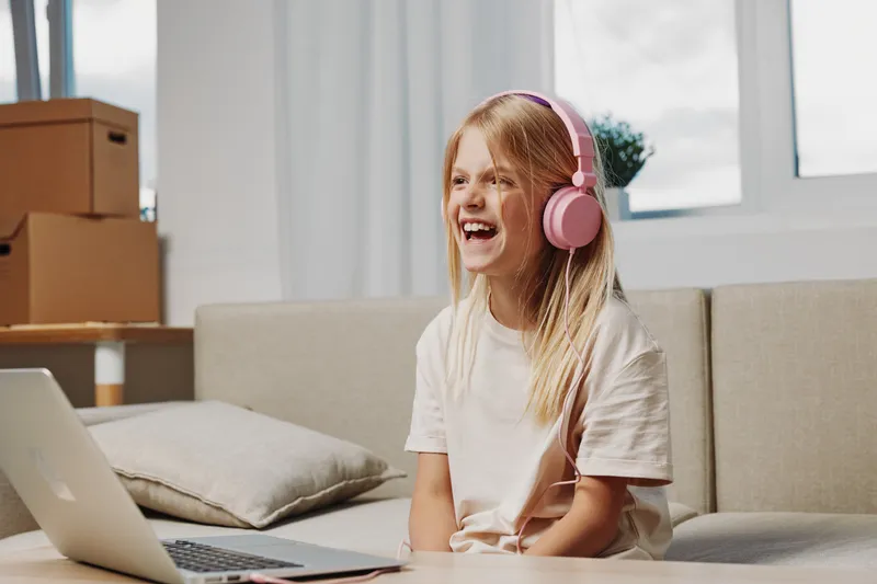 Happy blonde girl with pink headphones laughs while sitting on couch with laptop nearby in bright room. Concept of remote learning, entertainment, and childhood joy.