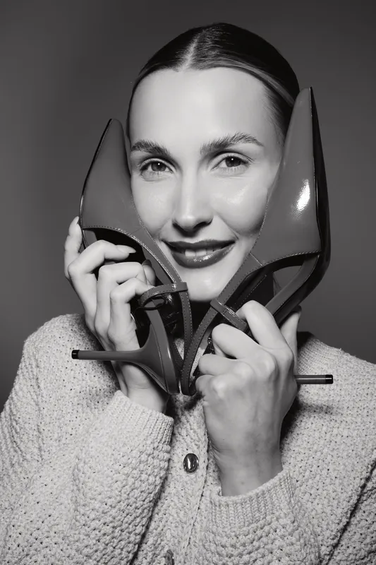 Portrait of a smiling Caucasian woman with fair skin holding red high heels near her face wearing a cozy sweater in studio. Concept of fashion, beauty, and footwear.