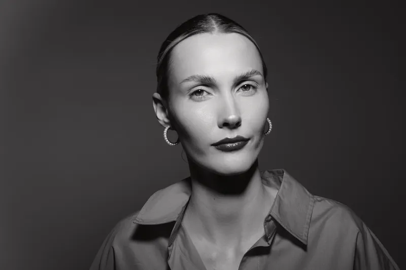 Serious white woman with fair skin and slicked back hair poses in studio. Black and white portrait with soft lighting highlights facial features and minimalistic style.