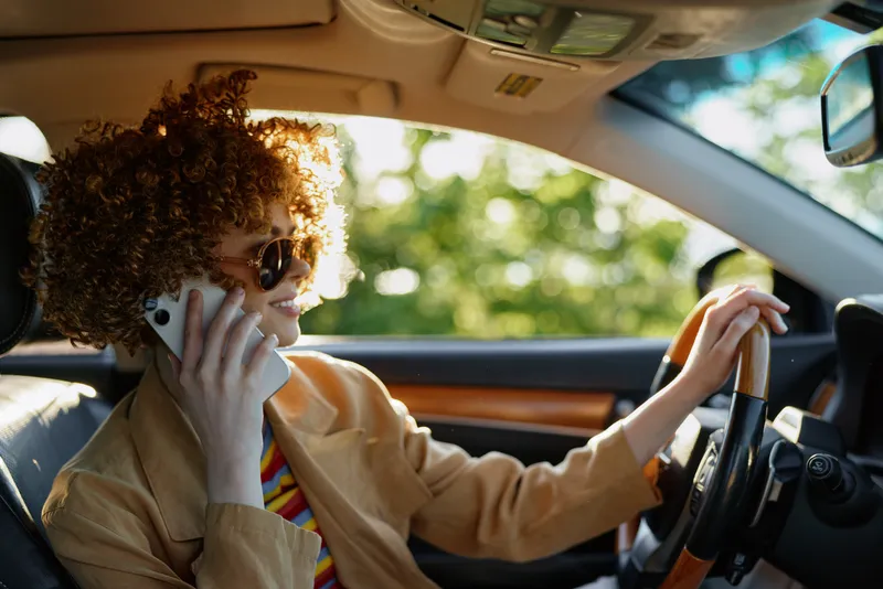 Smiling woman with curly hair driving a car, talking on a phone. Warm retro film color palette on a sunlit interior, conveying lifestyle, travel, and everyday optimism.