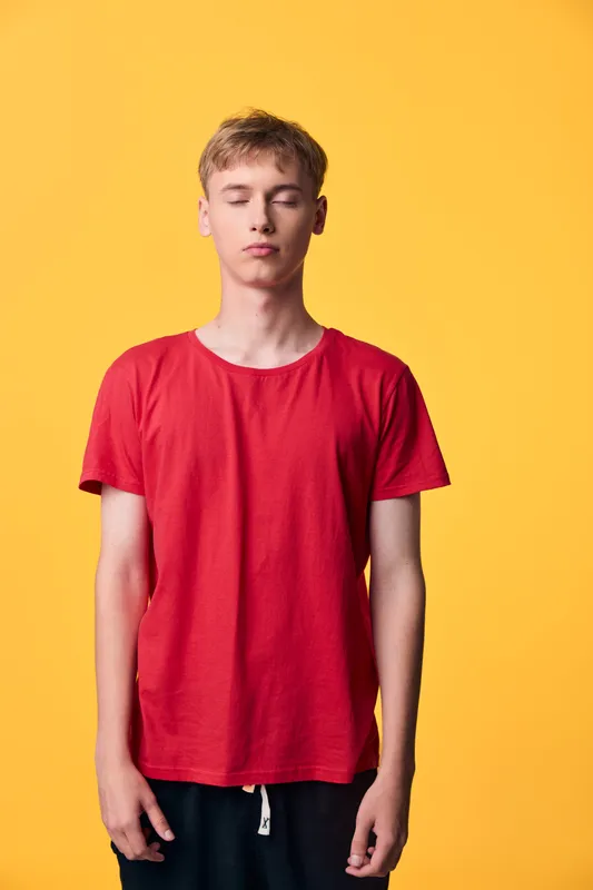 Red shirted young man stands with eyes closed in a bright yellow studio, conveying calm casual fashion in a minimalist studio setting for lifestyle and apparel catalogs