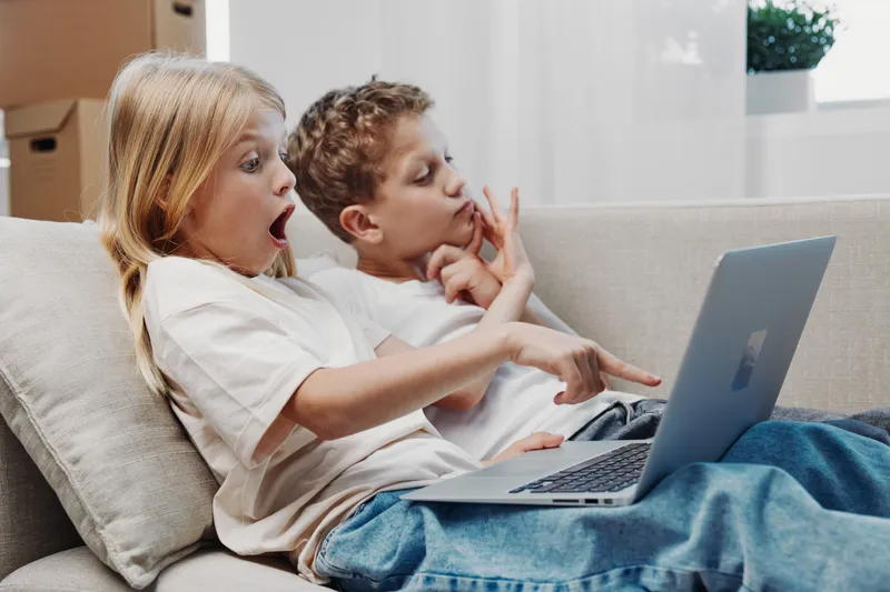 Two surprised children sitting on a sofa looking at laptop screen, pointing fingers and reacting with wide eyes and open mouth. Concept of curiosity, technology use, and childhood entertainment.