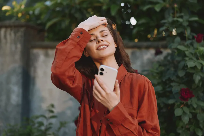 A young woman in an orange blouse smiles, shielding her eyes with one hand while holding a smartphone in a sunny garden with blooming roses, relaxed mood