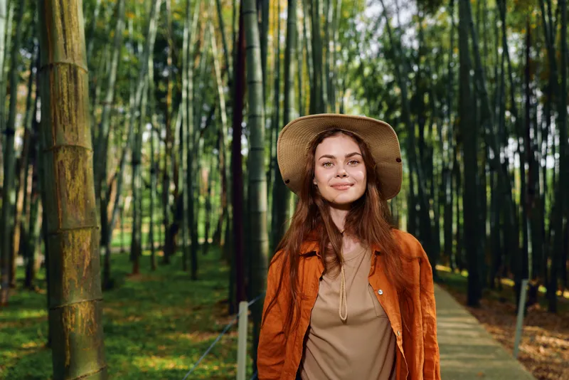 Woman in hat and portrait among bamboo forest on a wooden path, smile and relaxed nature scene with sunlight canopy and greenery, young female in casual jacket enjoying outdoors.