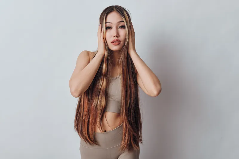 Young woman with long straight hair posing with hands on face, wearing beige crop top and pants against plain light background. Beauty, fashion and healthy hairstyle concept.