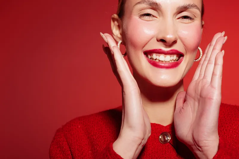 Smiling woman with red lipstick and earrings poses with hands near face on red background. Close-up portrait with bright makeup and joyful expression. Concept of beauty and happiness.