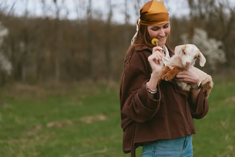 Woman outdoors with a goat kid, wearing a brown sweater and orange hat, offers a flower to the playful animal, showing warm affection and rustic lifestyle in a grassy field.
