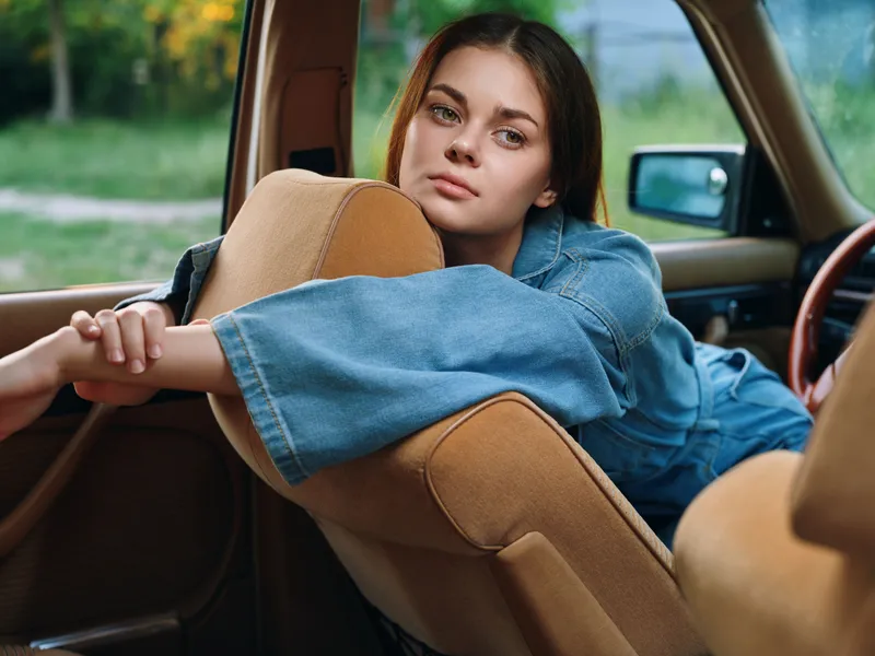 Thoughtful young woman in denim jacket relaxing in vintage car, emanating a serene and nostalgic mood, surrounded by lush greenery.