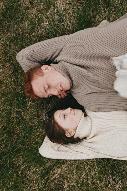 Relaxed couple lying on a grassy field, heads resting on soft blankets, sharing a quiet moment with a friendly dog nearby, evoking cozy outdoor lifestyle and gentle affection.