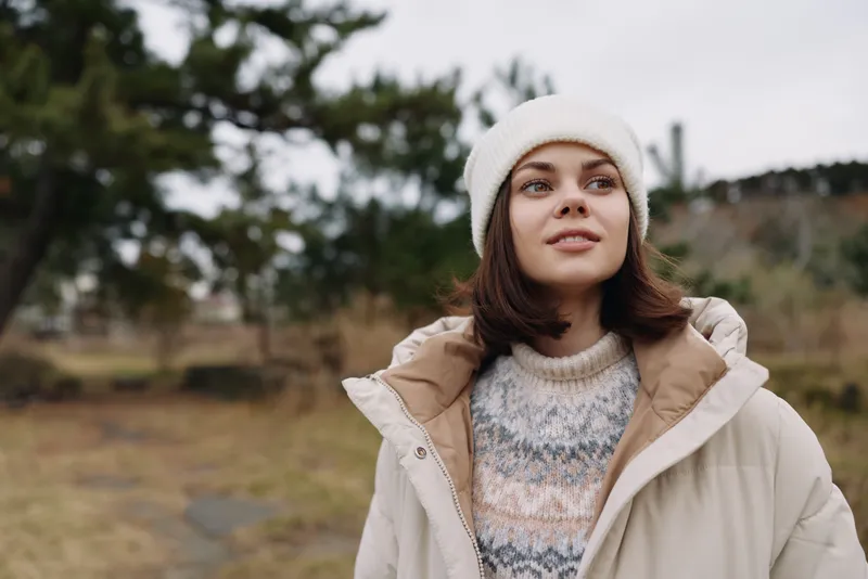 Woman in a warm winter coat and knit sweater outdoors, wearing a beanie and scarf, gazing upward with a calm expression in a wide natural park setting, framed by distant trees and cloudy sky