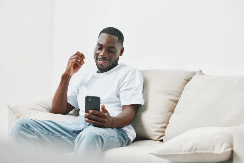 Smiling young Black man enjoying his smartphone while sitting on a cozy couch, dressed casually in a white t shirt and jeans, surrounded by a minimalist decor