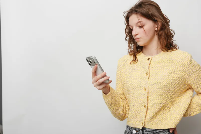 Young woman with curly hair wearing yellow knitted sweater looks at smartphone with focused expression. Isolated on light background. Concept of communication, technology, and casual style.