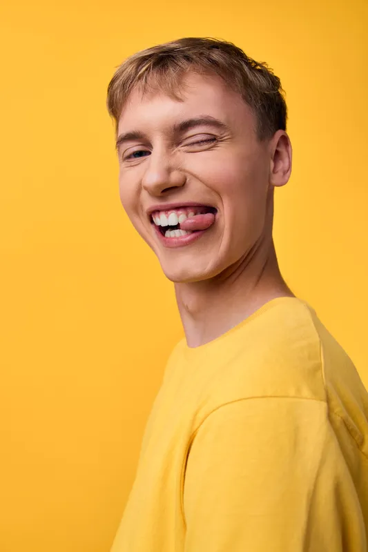 Smiling young man with tongue out in a bright yellow shirt against a vibrant yellow background, a playful expression conveying humor, energy, casual fashion style