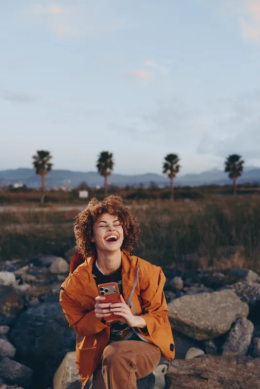 Happy woman lifestyle outdoor, smiling while holding smartphone, curly hair visible, wearing orange jacket, seated on rocks in palm landscape at sunset, candid travel vibe, fresh air today