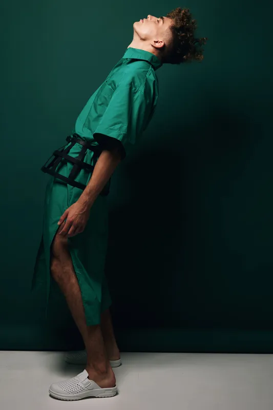 Young man in green outfit posing with head tilted back and bent body against dark background. Fashion model wearing casual clothes and white shoes in studio setting.