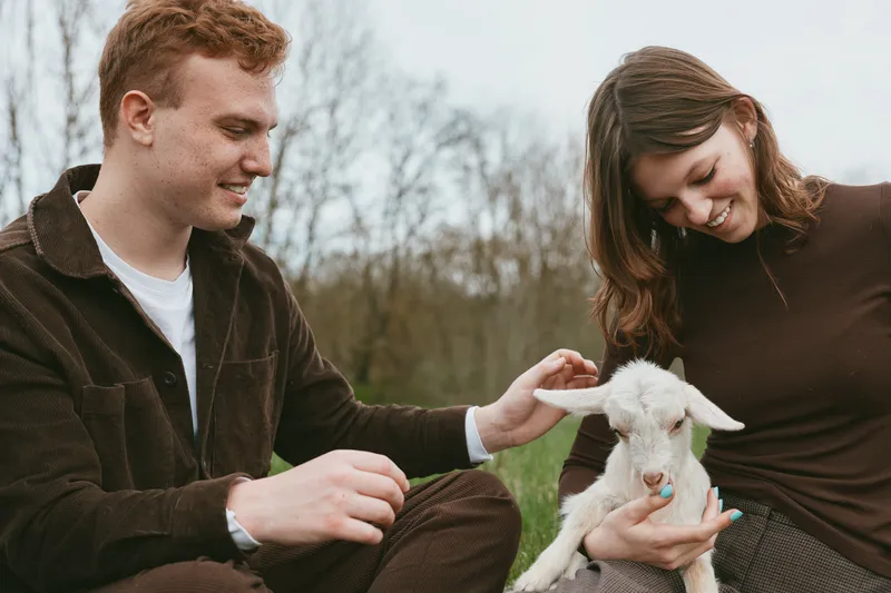 Young man and woman sitting outdoors gently pet a white baby goat in a natural setting during a calm autumn day, enjoying a peaceful moment with the animal.