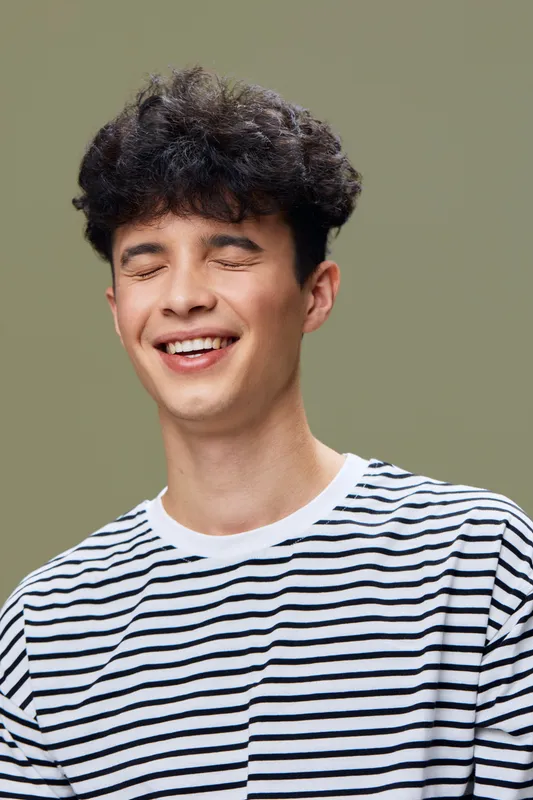 Young man with curly hair smiling with eyes closed wearing a striped shirt, joyful expression on green background, casual and natural mood portrait.