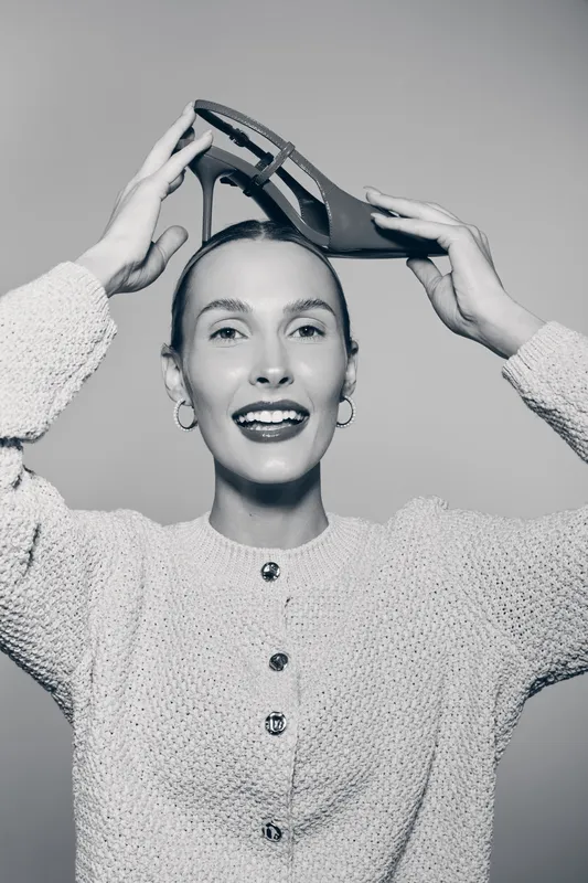 Smiling Caucasian woman with fair skin holding high heel shoe above her head wearing knitted sweater. Concept for fashion, style, footwear, and joyful mood in studio portrait.