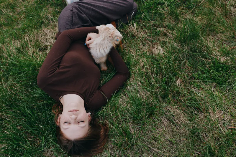 Relaxed lifestyle photograph of a woman lying on grass, holding a goat kid, showcasing calm nature and outdoor bond between human and animal