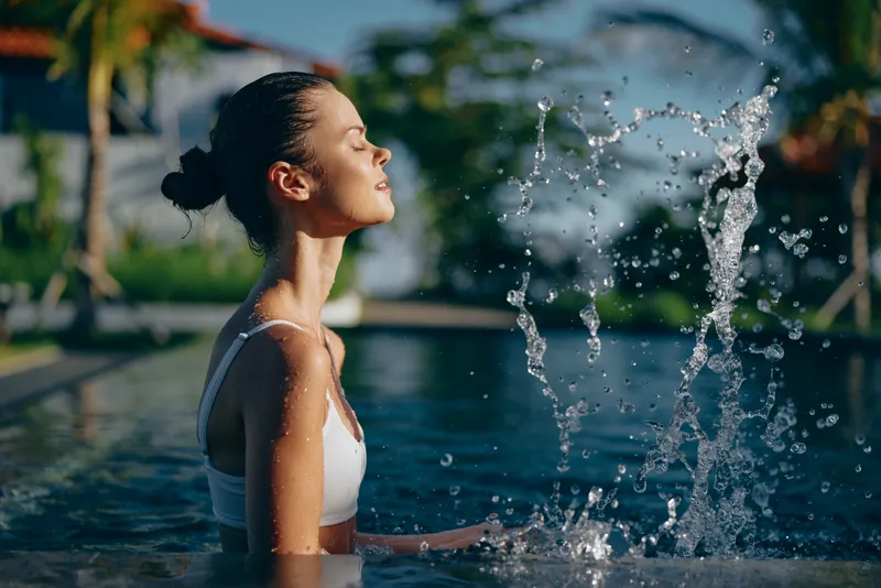 Young woman with brunette hair enjoys a peaceful moment standing in a swimming pool as water splashes nearby under warm sunlight.