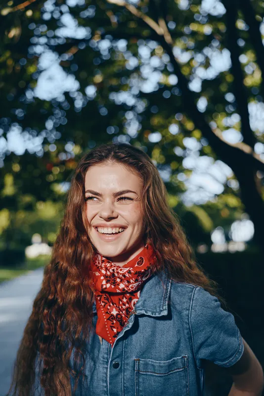 smiling woman with long curly hair wearing red scarf and denim jacket outdoors under green trees in sunny daylight expressing happiness and joy