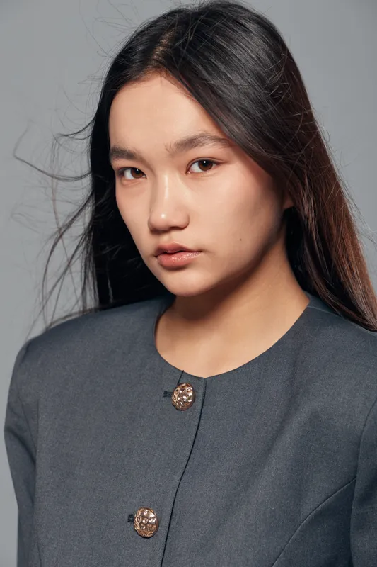 Portrait of young woman with long dark hair wearing gray blazer with decorative buttons, looking at camera with neutral expression in studio closeup with plain background.