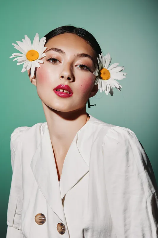 Portrait of a young woman with vibrant makeup and fresh daisies in her hair poses confidently against a green background. Creative beauty and fashion concept with elegant style.