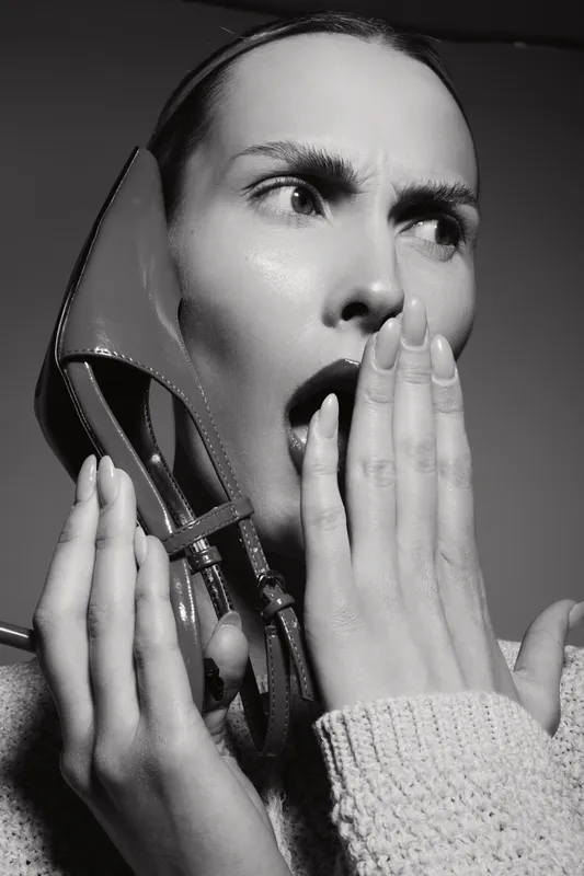 Surprised Caucasian woman with pale skin holds high heel shoe near face, covered mouth with hand. Concept of fashion shock and emotions in studio.