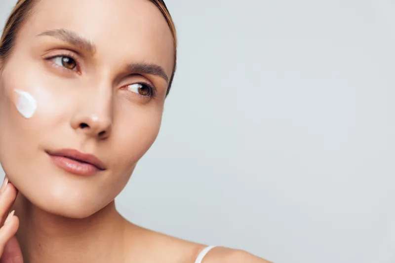 A young woman applies moisturizing cream on her cheek, showing clear skin and natural beauty against a plain light background. Concept for skincare, health, and self care routines.