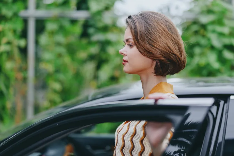 Profile view of a woman stepping from a sleek car, wearing a striped knit sweater, captured in soft natural daylight.