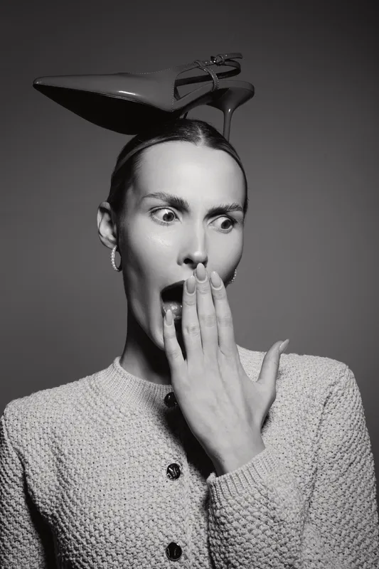 Surprised slender Caucasian woman with fair skin and dark hair holds hand near mouth, wearing high heel shoe on head in black and white portrait studio.