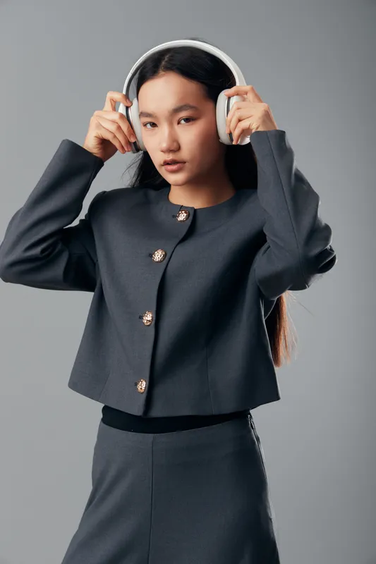 Young woman wearing grey two piece suit adjusts wireless headphones for music or podcast listening in studio. Elegant casual outfit with long hair and focused expression on face.