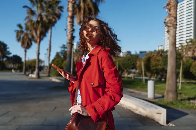 Young woman in a red trench coat walks outdoors on a sunny day, holding a smartphone and glancing to the side with a confident, relaxed vibe.
