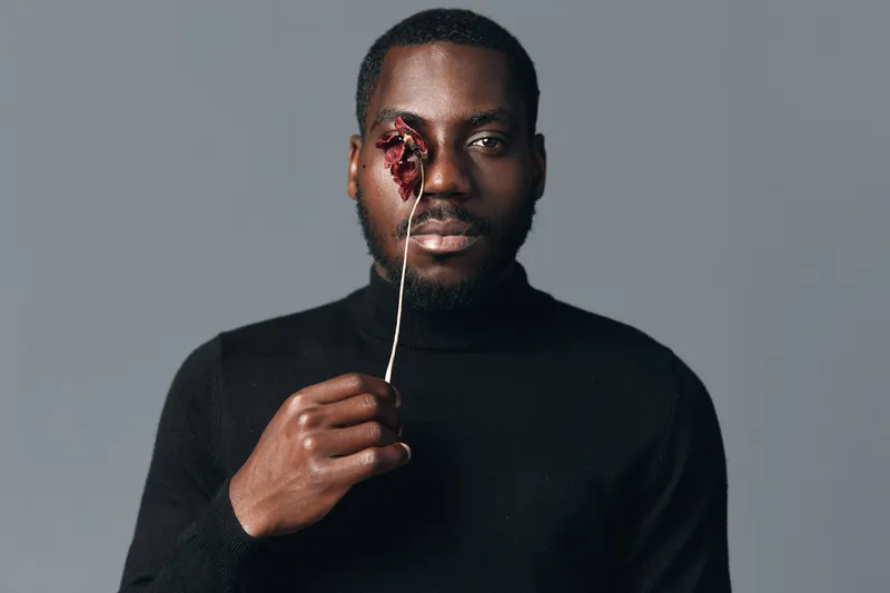 Black man with dark skin holding dried flower in front of eye in creative visual designer style. Portrait with serious expression and minimalistic gray background for artistic concept.