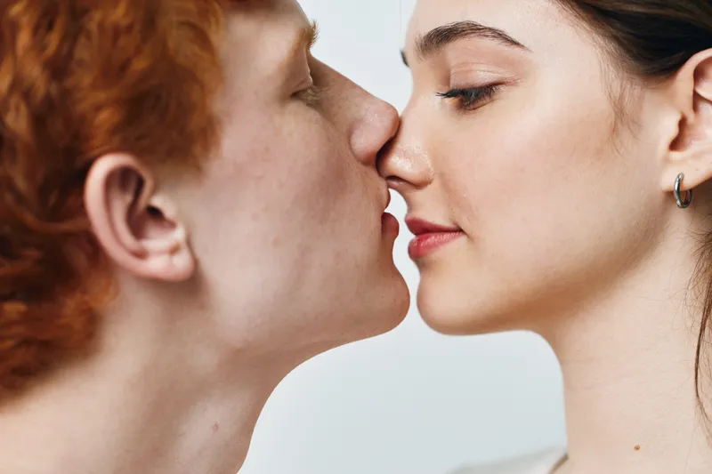 Couple with red hair and dark hair close together touching faces for intimacy love and affection in studio with neutral background. Young man and woman with eyes closed.
