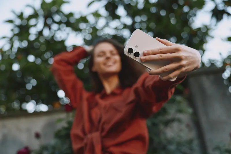 A woman in a red shirt takes a selfie outdoors, smiling with a phone held high. A candid lifestyle portrait capturing casual confidence, joy, and everyday moments in daylight