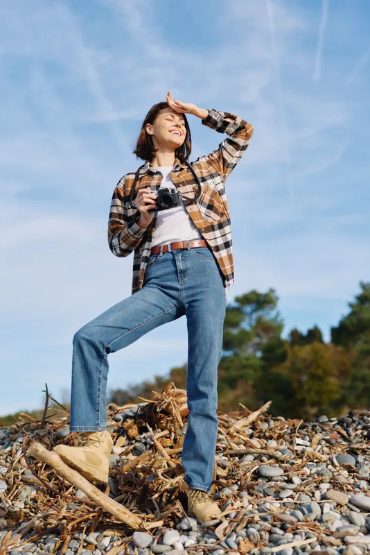 Woman with camera in outdoor rocky terrain, wearing a plaid shirt, jeans and boots, looking toward the horizon under a clear blue sky, capturing candid travel moments.
